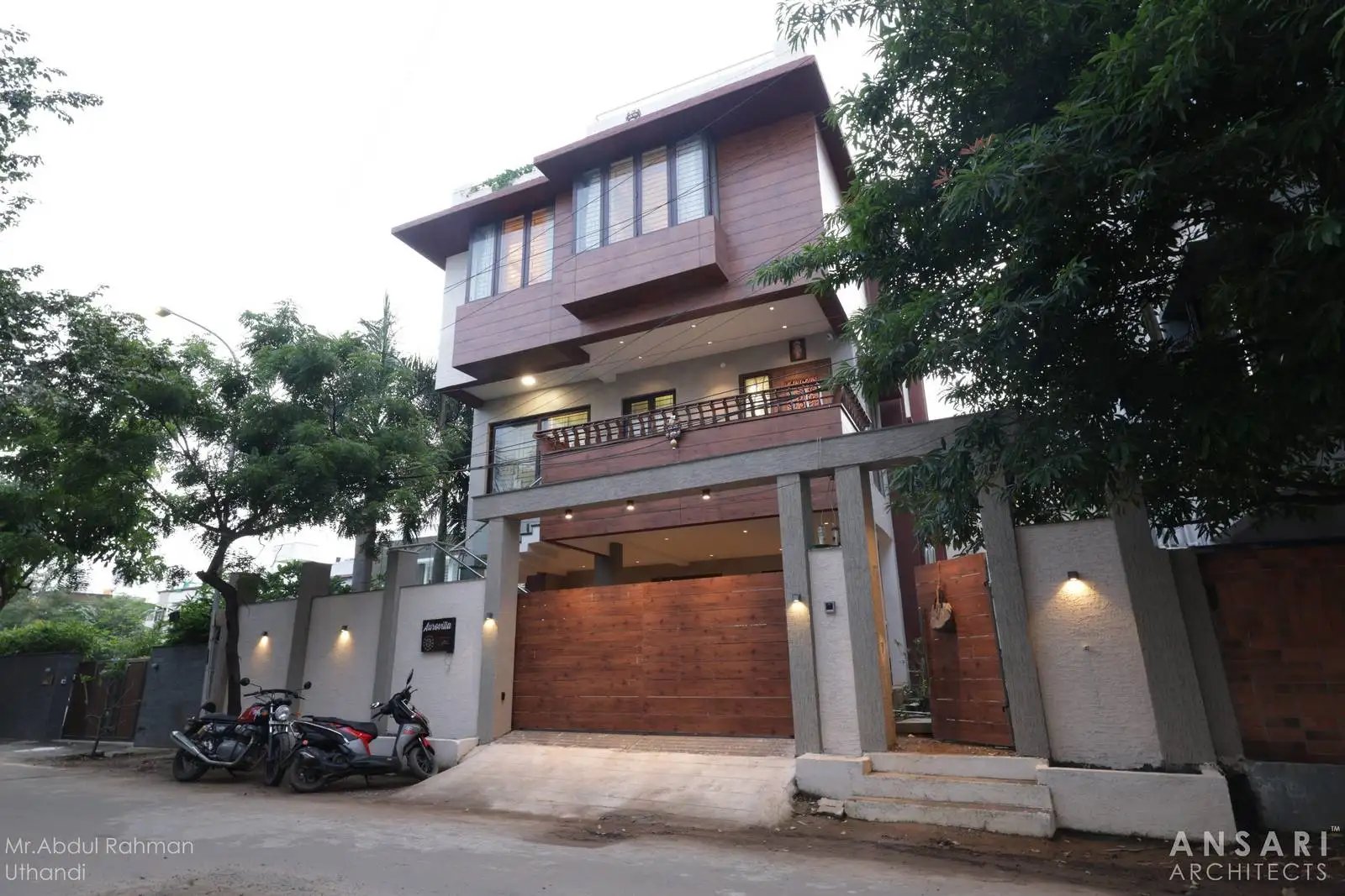 Modern two-story residential building with a prominent wooden garage door and balcony, surrounded by trees and a light-colored concrete fence with mounted lights, featuring motorcycles parked on the street outside.