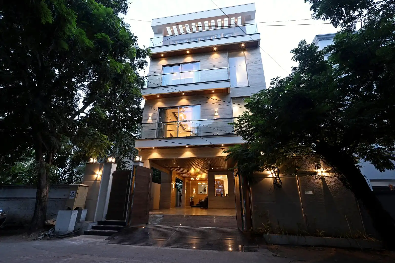 Modern four-story house with large glass windows and balcony railings, warmly lit exterior at dusk, surrounded by trees and open wooden gates at the entrance.