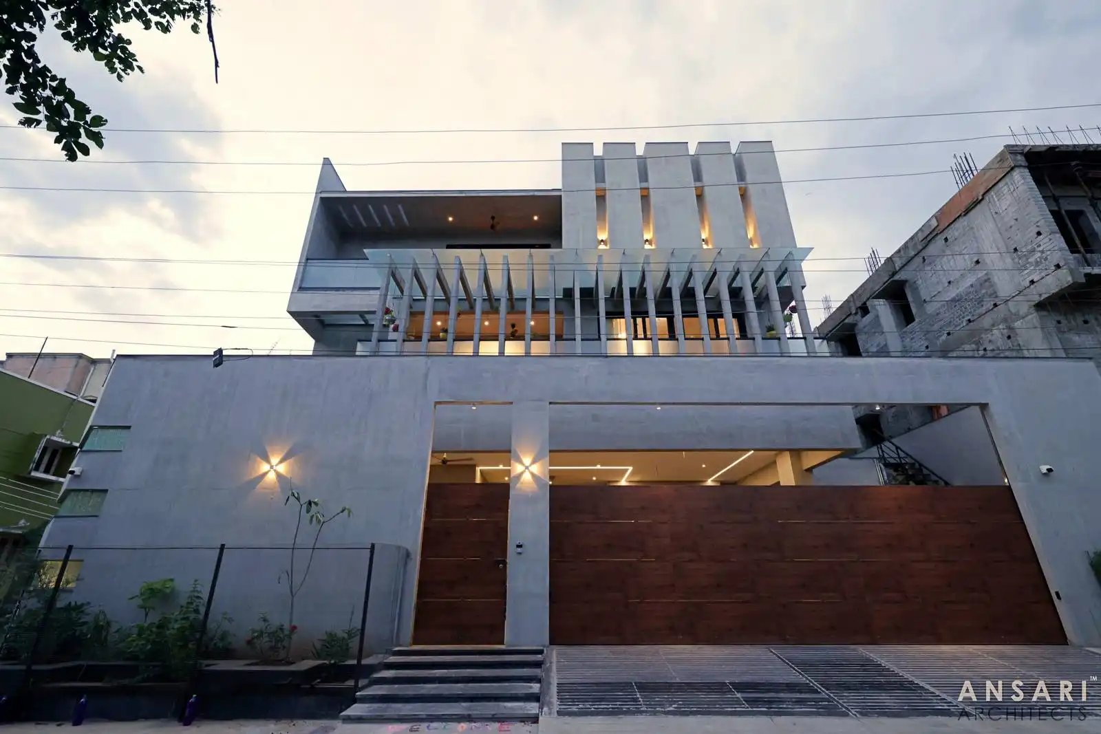 Modern multi-story house with concrete facade, vertical white slats on the balcony, warm interior lighting, large wooden gate, and small garden area in front during dusk.