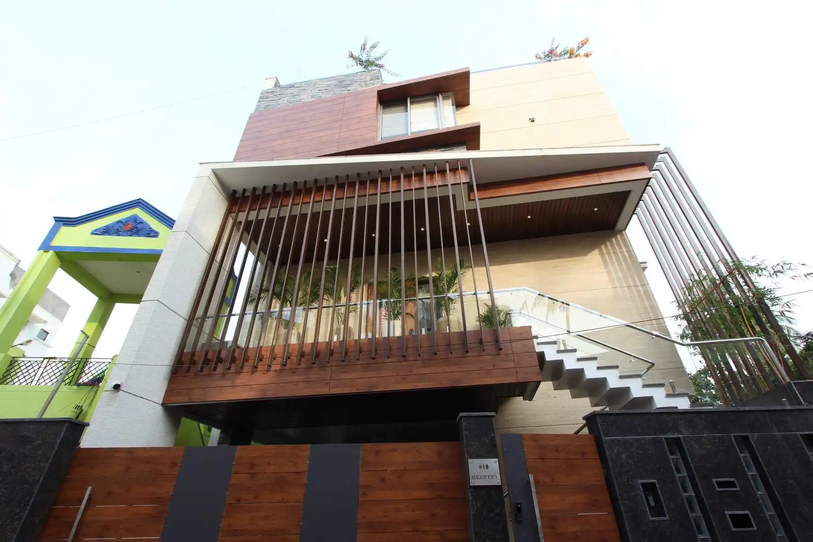 Modern multi-story house with wooden and stone facade, glass balcony railing, vertical wooden slats, an external staircase, and adjacent green building entrance.