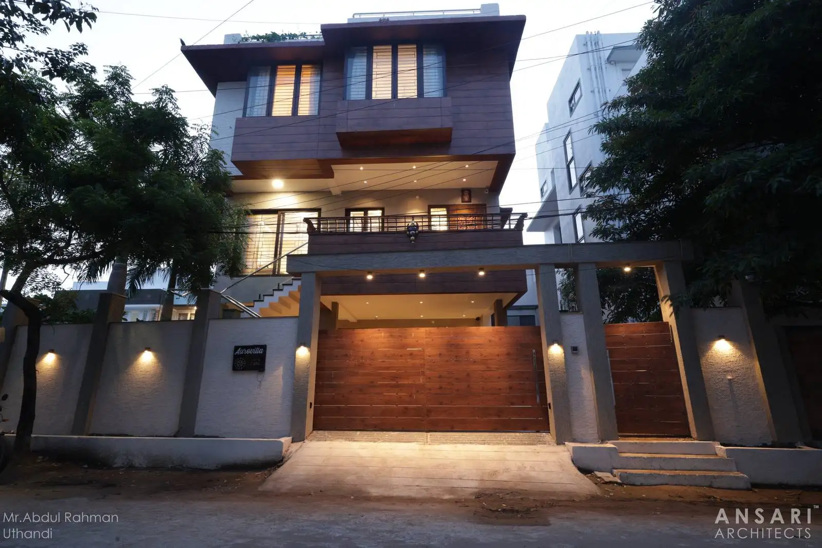 The entrance gate combines warm wooden planks with light textured boundary walls, creating a welcoming yet secure look. Subtle integrated lighting enhances textures and highlights the design, making the entryway elegant by day and striking by night.
