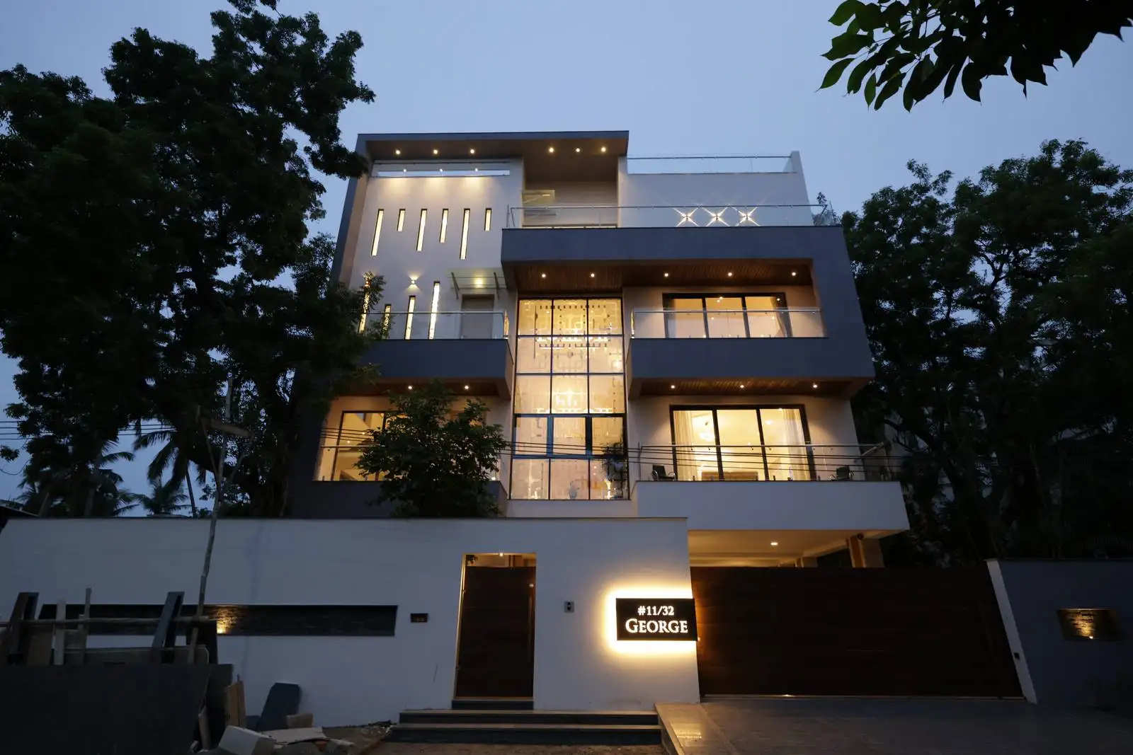 Modern three-story house at dusk with large windows illuminated from inside, white and gray exterior, surrounded by trees, and a glowing sign near the entrance that reads #11/32 GEORGE.