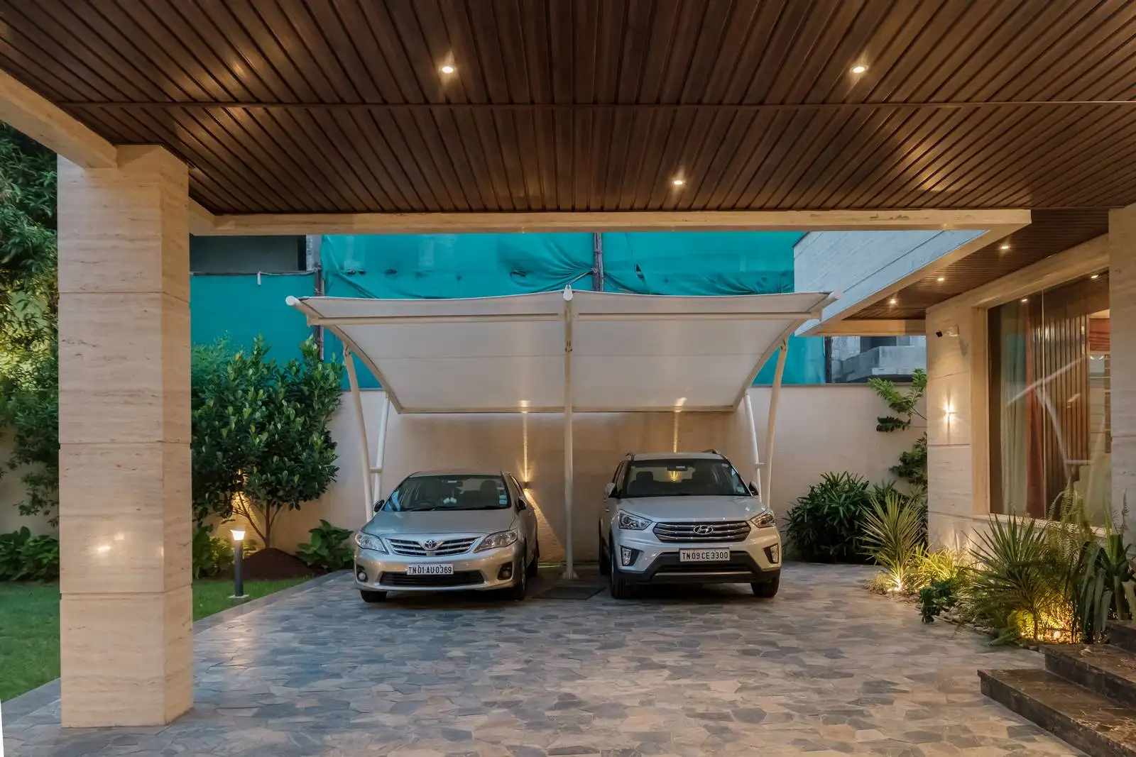Covered driveway featuring a sleek tensile fabric canopy and natural stone flooring. Side-by-side parking is integrated with soft landscaping and warm ambient lighting, balancing functionality with refined aesthetics.
