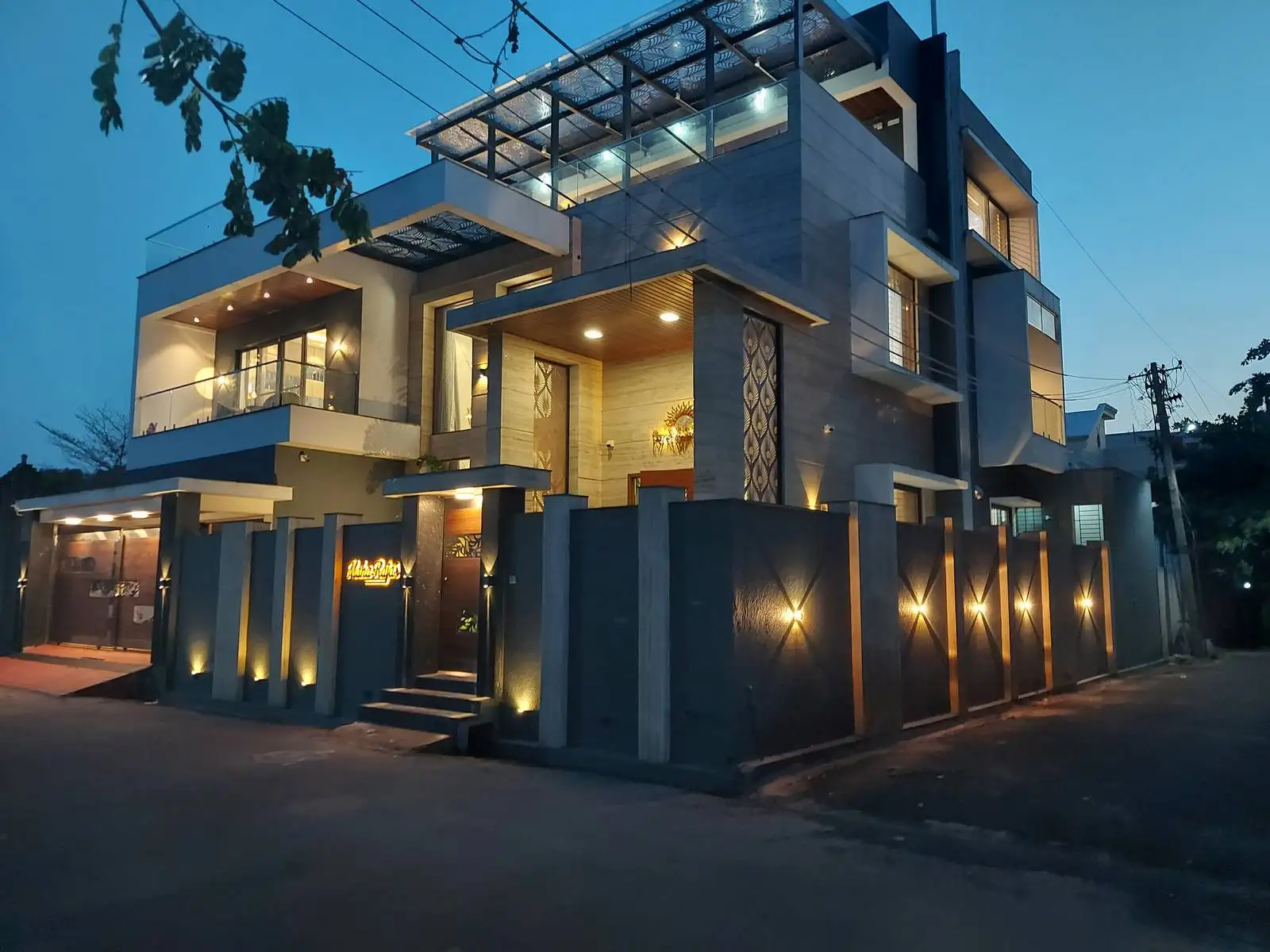 Modern two-story house illuminated with warm exterior and interior lights at dusk, featuring a gated fence, glass balcony, and contemporary architectural design.