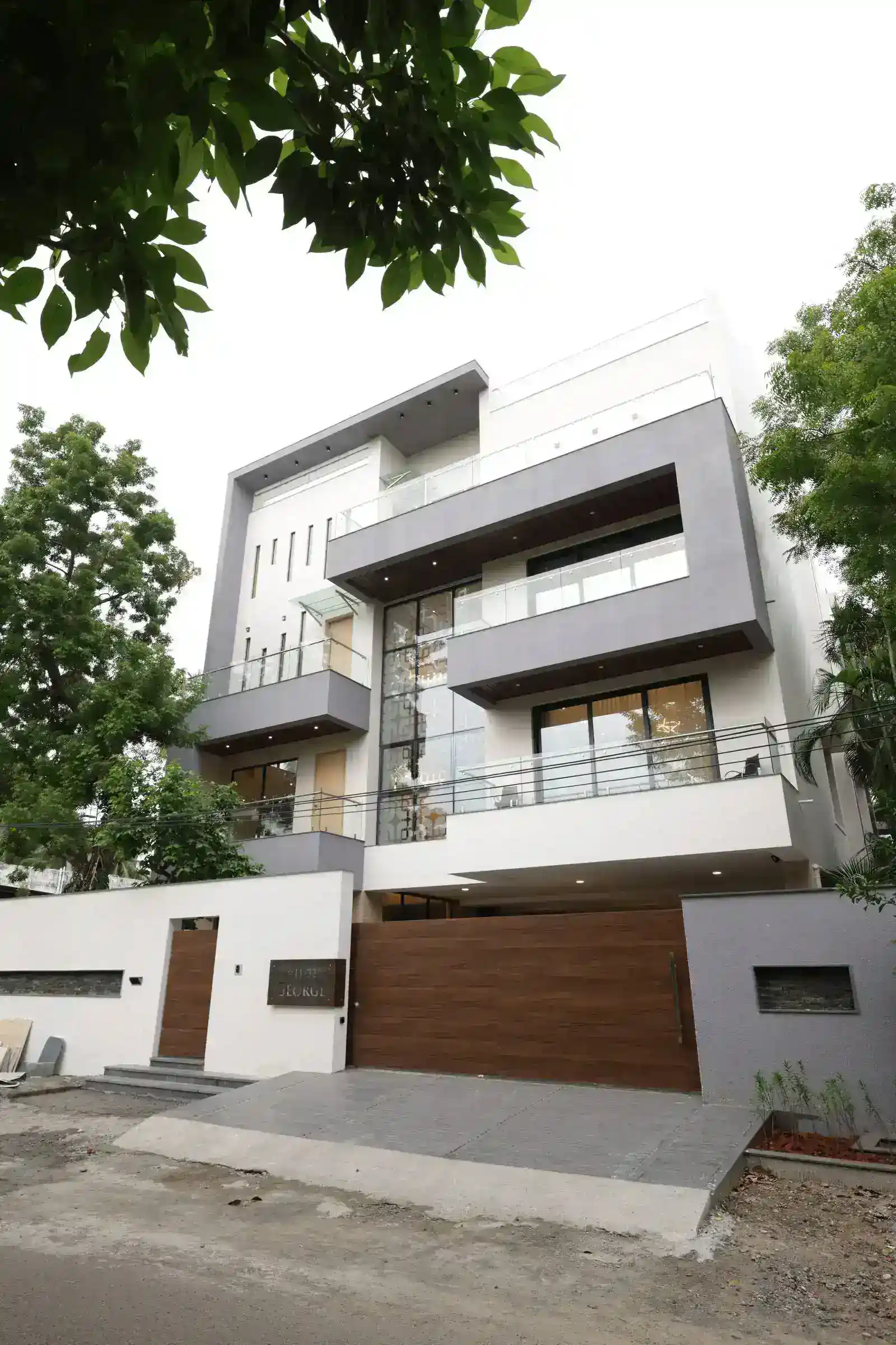 The main gate and pedestrian entry are integrated into a clean, white boundary wall, providing privacy and security.Glass railings and large windows on the upper floors add transparency and a modern touch, allowing natural light to flood the interiors.
