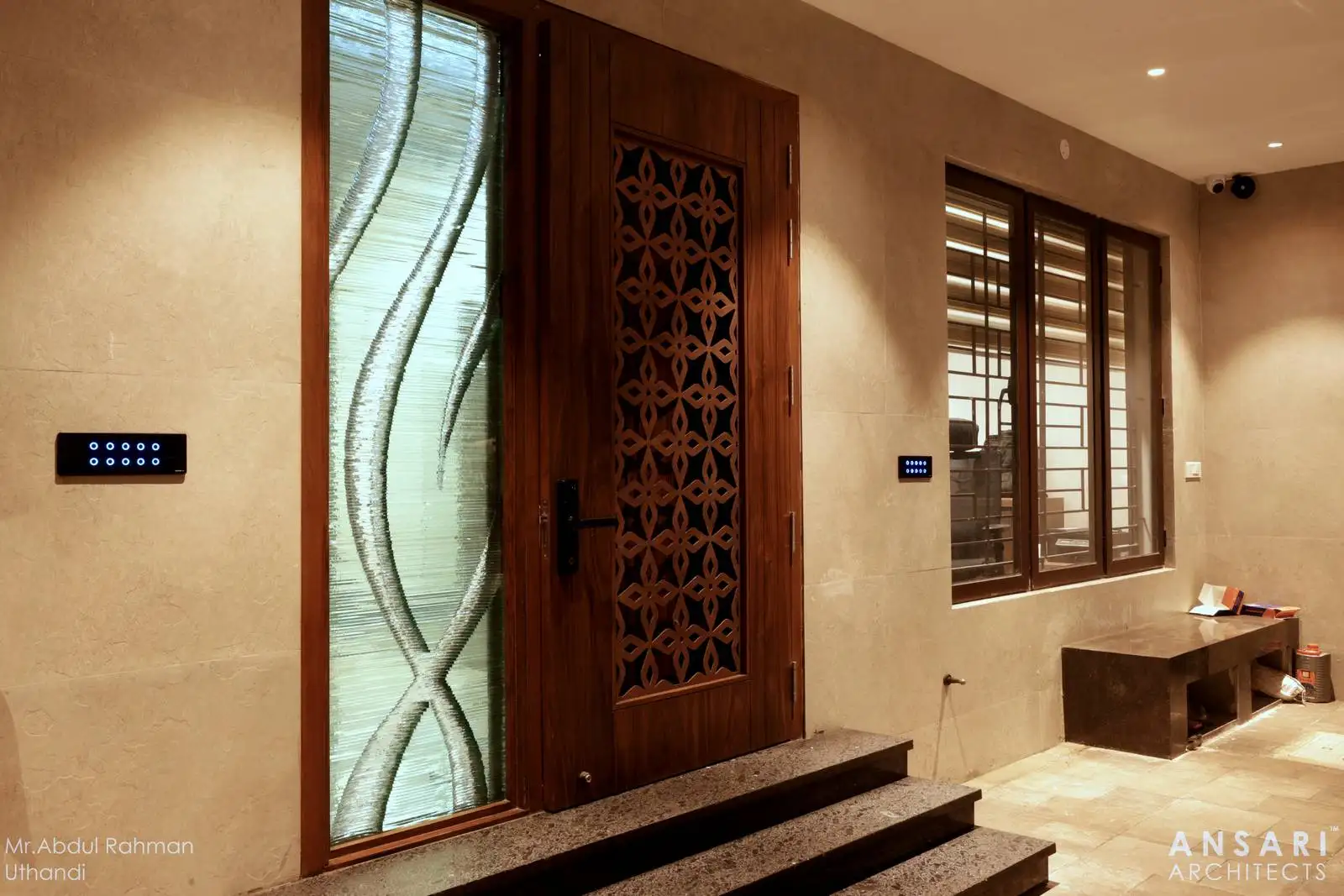 The main entrance features a solid wooden door with decorative panels and a side glass panel with artistic frosted patterns, adding elegance and natural light.