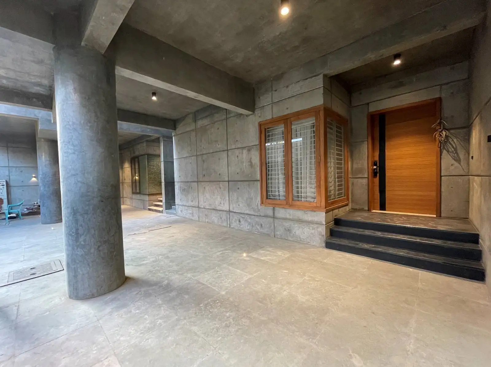 The car parking and verandah combine brutalist concrete with functional design, featuring clean structures, minimalist detailing, and durable stone flooring. Timber accents and soft lighting add warmth, creating a welcoming transitional space.