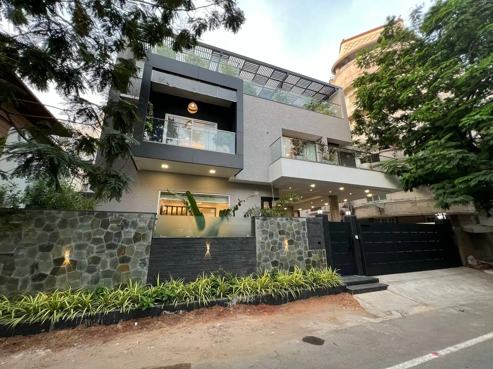 Modern two-story house with a stone and glass front fence, large black gate, and balconies featuring glass railings and plants, surrounded by tall trees on both sides. The Bianco Villa