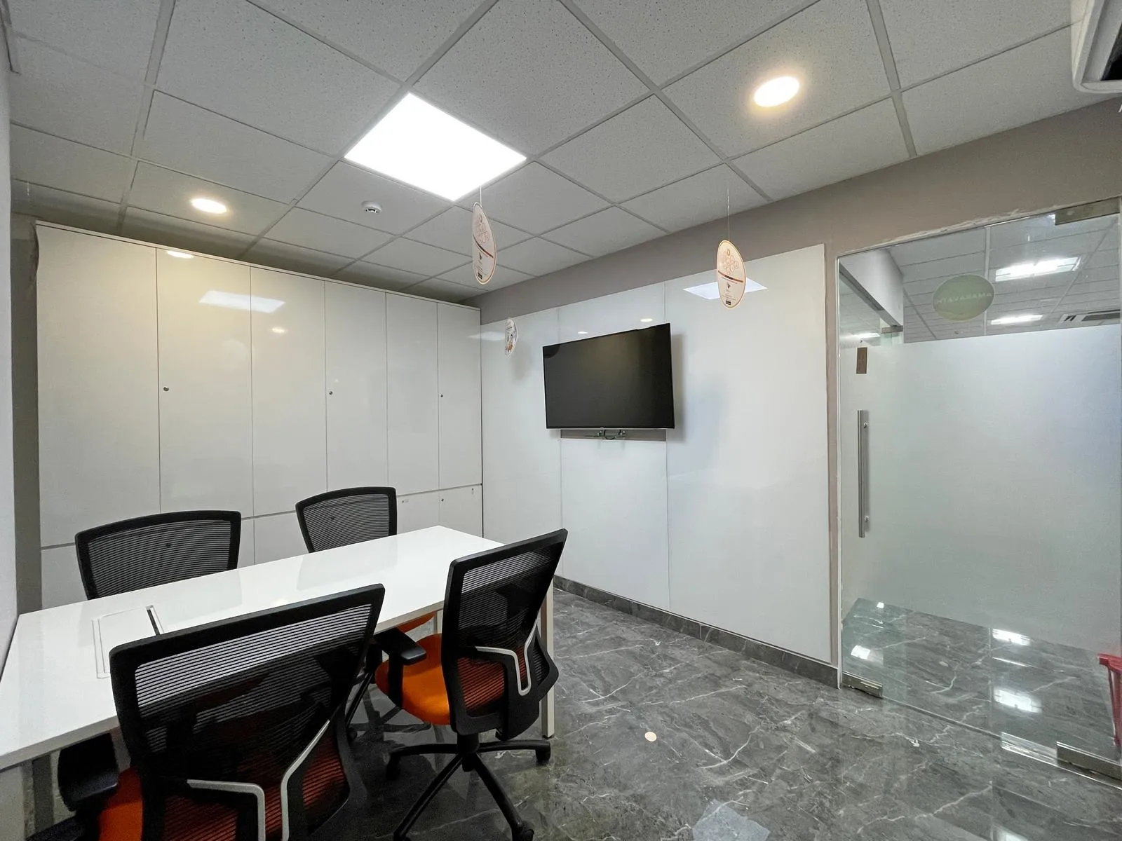 Meeting Room 2 features a modern four-seater layout with ergonomic chairs, a digital display, and natural light, designed for productive, technology-enabled collaboration. Sleek white finishes, marble-effect flooring, and integrated lighting create a professional and contemporary corporate space.