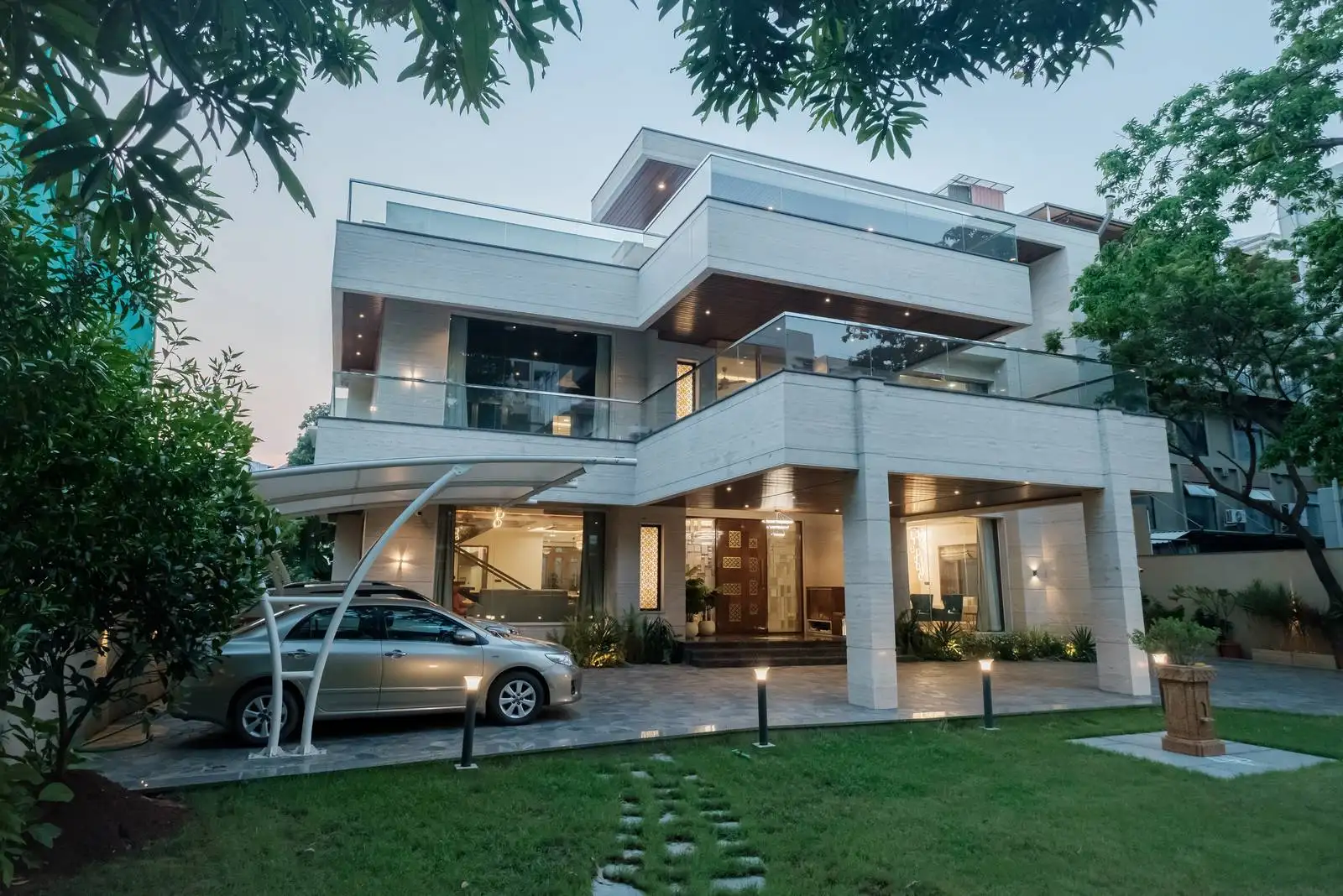 Home features a clean white exterior, expansive glass balconies, and a sculptural curved metal carport over a stone driveway—combining minimalist design with functional elegance.
