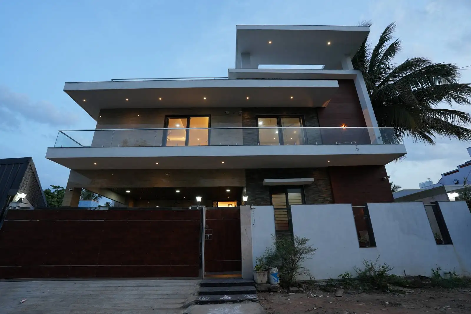 Modern two-story house with illuminated windows and balcony at dusk, featuring a large dark wooden front gate, white boundary wall, and a tall palm tree on the right side.