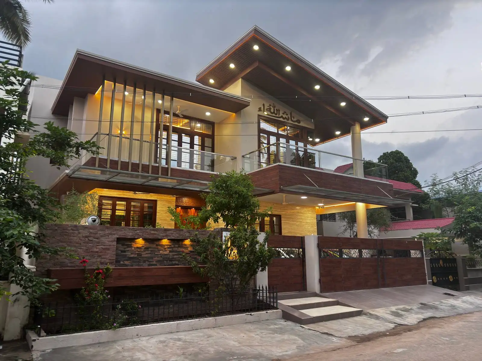 the infinity villa Modern two-story house with a slanted roof, illuminated by outdoor lights, with large glass doors and windows, a wooden and stone facade, a gated entrance, and some trees and greenery in front under a cloudy sky.