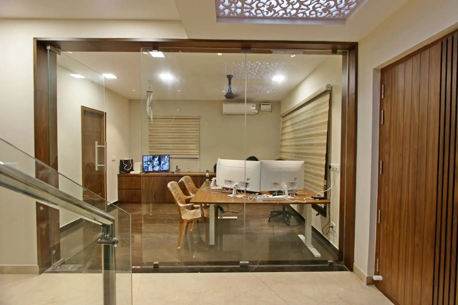 The office features a full-height glass partition with wooden framing, offering transparency and acoustic privacy. A spacious wooden desk with dual workstations ensures functionality and modern elegance.