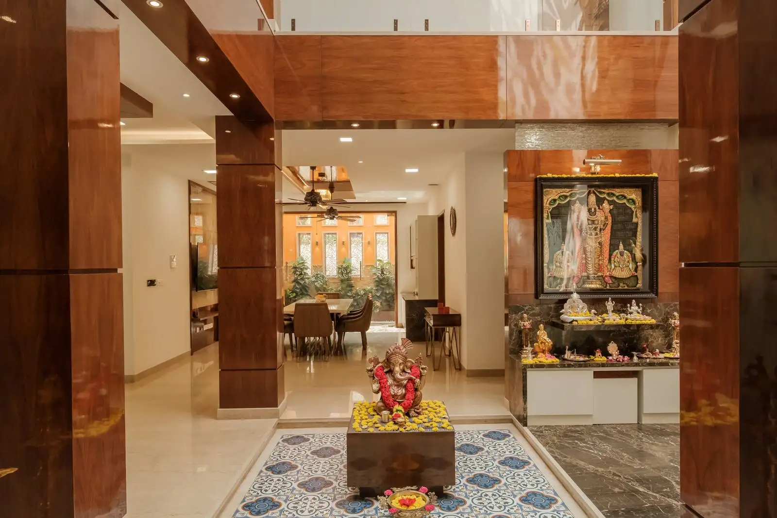 Pooja area with a central Lord Ganesha statue on a patterned tile platform, framed by polished wooden pillars and adjacent to a dining space, blending spiritual and functional zones with elegant detailing.
