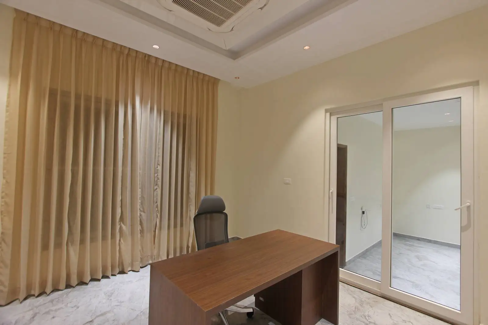 Minimalist office with a wood desk, ergonomic chair, and large windows with curtains. Recessed lighting and glass partition enhance comfort and privacy.