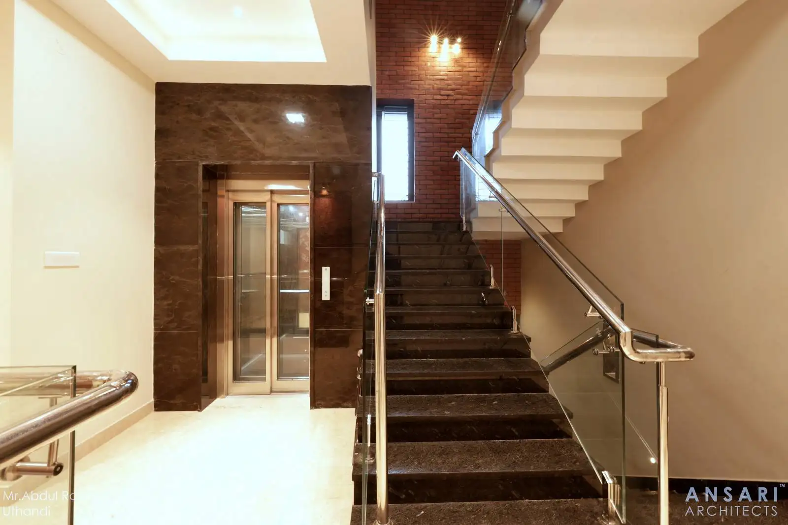 An indoor staircase with dark stone steps and a glass railing seamlessly connects to the second floor, positioned beside a sleek elevator with metallic doors framed by dark stone walls, all highlighted under bright ceiling lighting for a striking, contemporary look.