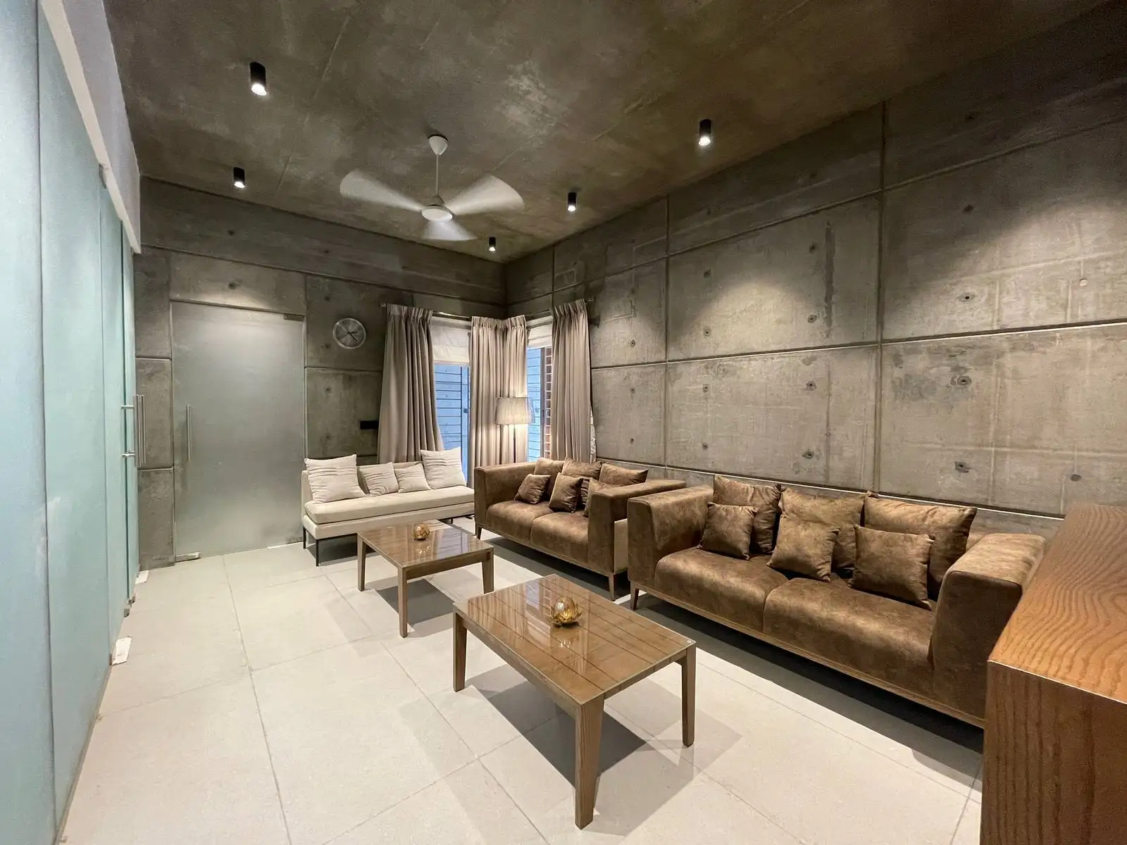 The lounge features concrete walls, grey vitrified flooring, brown suede seating, and solid wood tables for a warm, balanced look. Frosted glass partitions, ceiling spotlights, and neutral curtains enhance light, privacy, and depth.