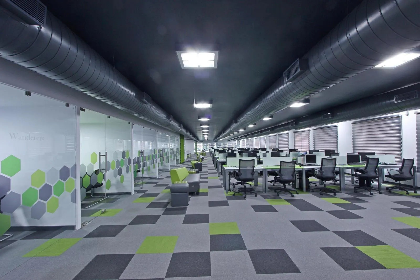 Contemporary workspace design with modular workstations, ergonomic chairs, and patterned carpet tiles in neutral and accent tones. Industrial-style black ceilings, glass partitions with custom graphics, and collaborative seating zones enhance functionality and aesthetics.