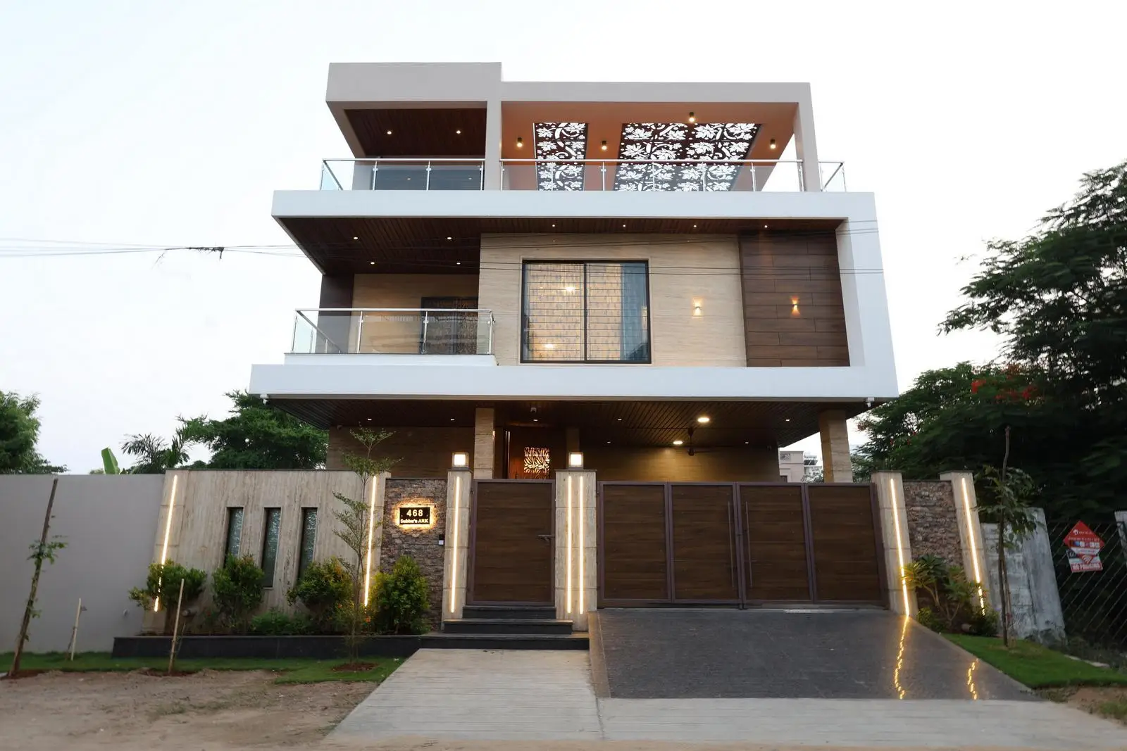 The modern exterior features a rectilinear form with strong horizontal lines, cantilevered balconies, and dynamic volumes. The facade combines white plaster, wood-textured cladding, glass railings, and stone accents for a warm, contemporary look. 