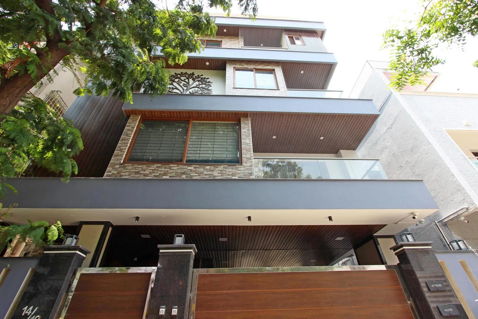 Modern multi-story house with a large wooden gate, stone and wood exterior accents, glass balcony railings, and greenery with trees surrounding the front area.