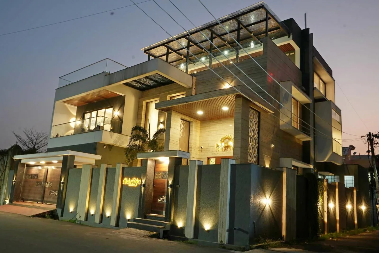 Modern multi-story house illuminated with warm lights at dusk, featuring large windows, a balcony with glass railing, and a decorative front entrance wall with vertical lights.