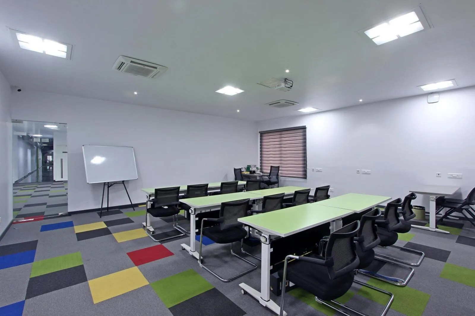 Contemporary training room design featuring modular lime green tables, ergonomic mesh seating, and multi-colored carpet tiles for dynamic spatial zoning. Matte white walls, dual-tone blinds, recessed LED lighting, and integrated air-conditioning ensure a bright, comfortable learning environment.