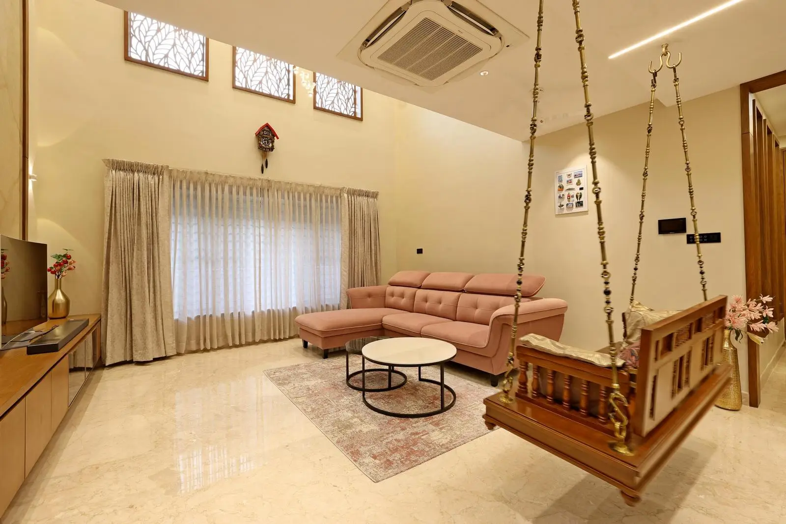 The double-height living area enhances openness and light, with tall windows emphasizing verticality. Neutral marble flooring, wooden furniture, and a pastel sofa create a balanced, warm palette. A custom swing, sleek furnishings, and minimal décor blend modern minimalism with subtle traditional character.