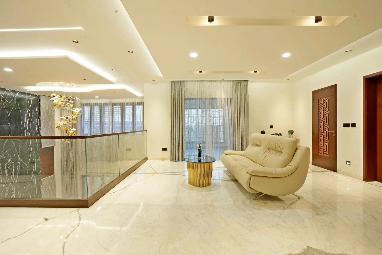 The first-floor 19'0" x 18'0" family hall overlooks the double-height living area below, creating openness and visual connectivity. White marble flooring, soft off-white walls, warm wooden accents, and a minimalist seating arrangement combine luxury, calmness, and sophisticated elegance.