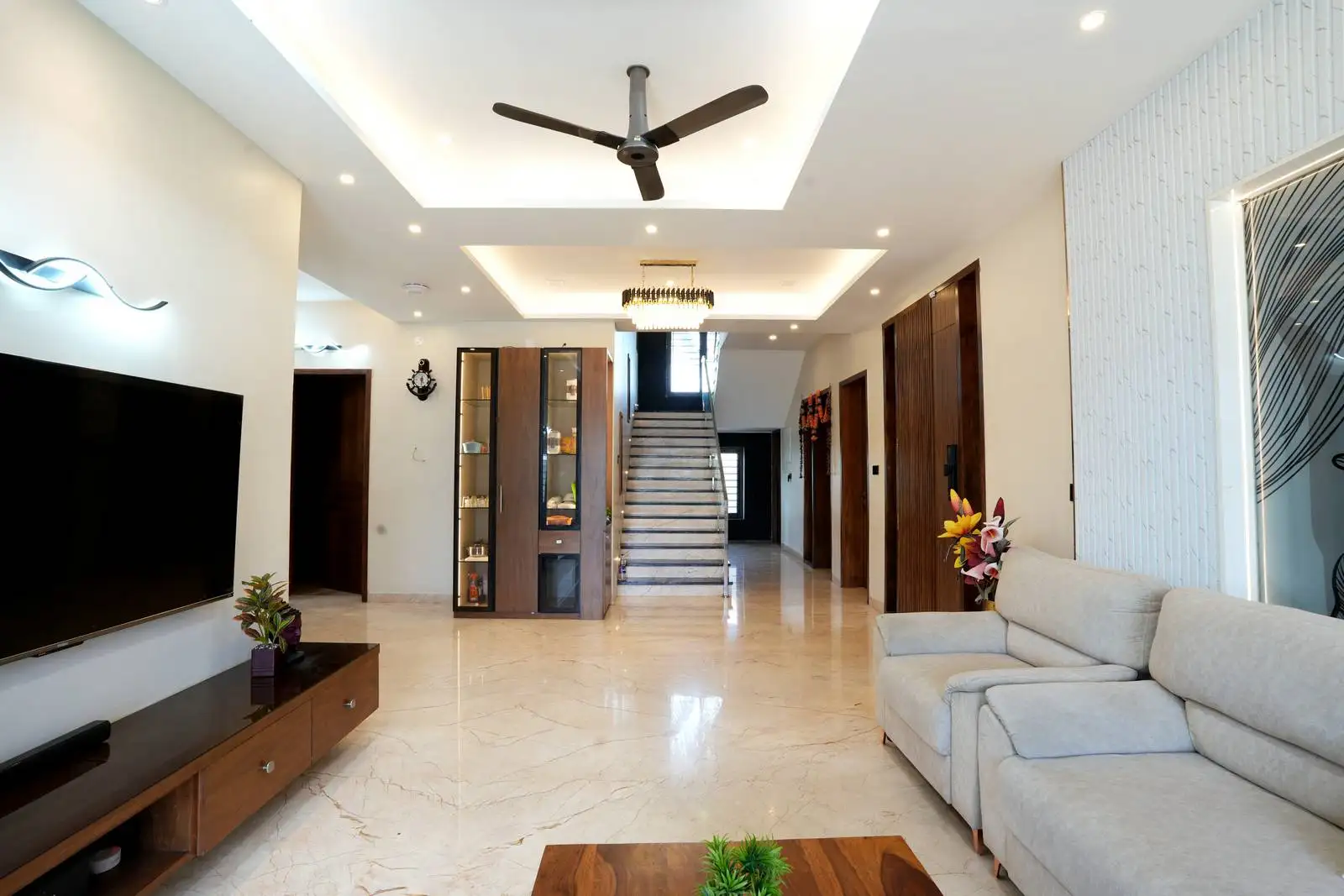 Living room with layered ceilings, integrated lighting, and a focal staircase. Warm wood accents and minimalist furniture enhance the open-plan layout and natural light.