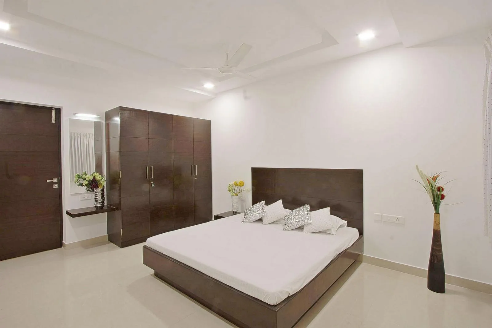 The bedroom pairs white walls and reflective floors with dark walnut furniture for a bright yet warm feel. Minimal furnishings, a floating dressing counter, and a low bed highlight openness, horizontal lines, and modern elegance.