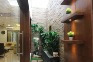 Indoor corridor with a stone-textured wall, green plants in planters, wooden shelves with small potted plants, glass door with stainless steel handle, and a glimpse into a living room with beige sofas.