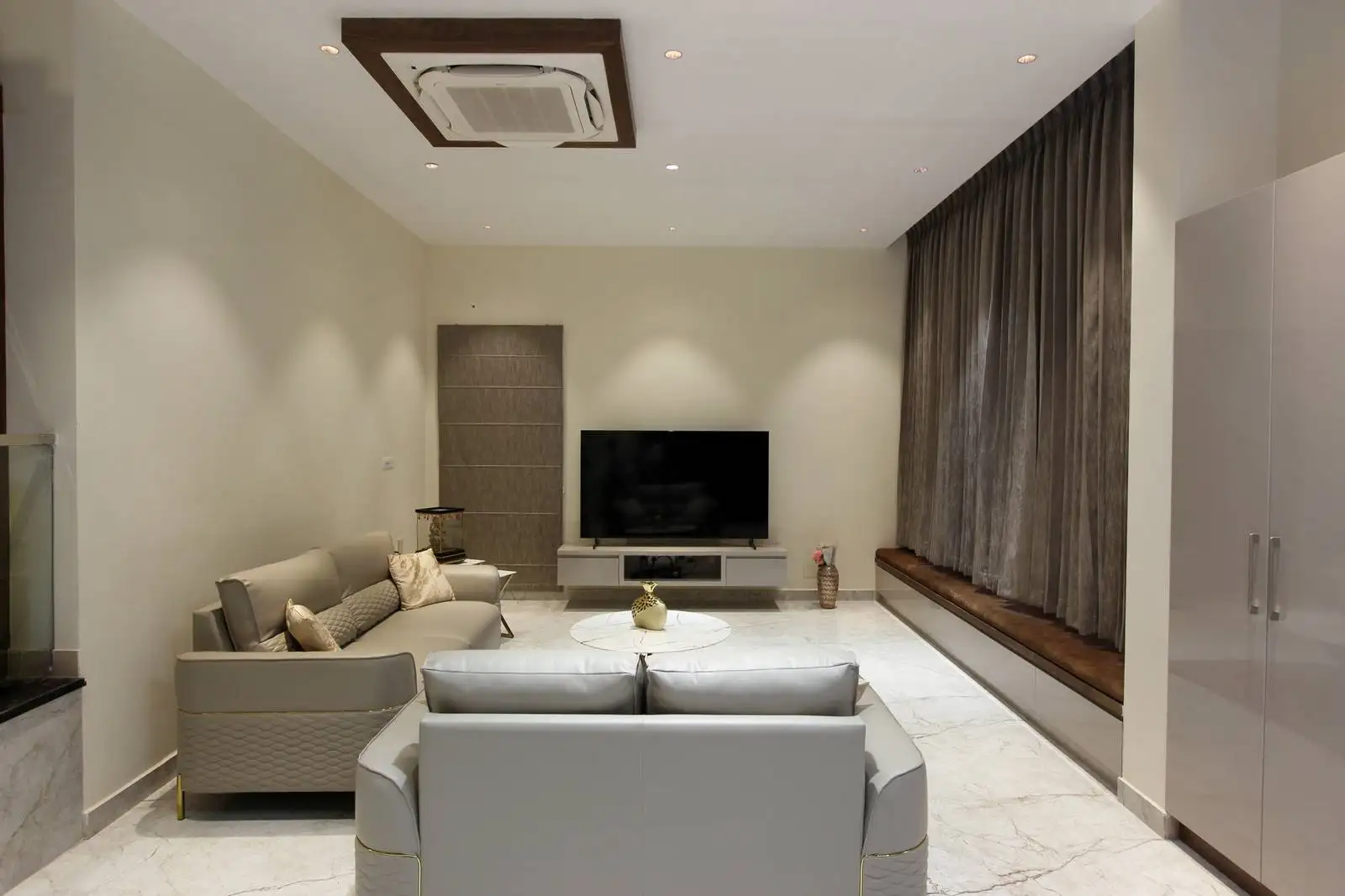 The living room includes large windows, built-in wooden seating, and light marble flooring that enhances brightness. Recessed lighting and concealed storage keep the space organized and inviting.