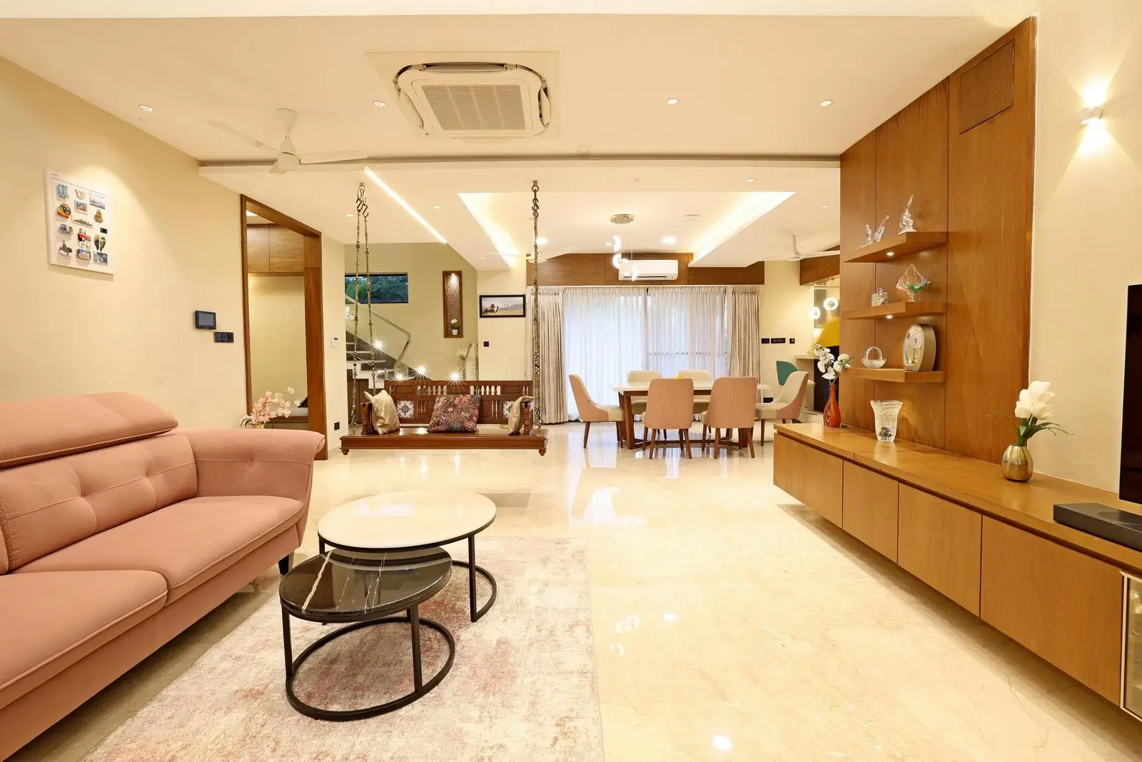 The open-plan layout seamlessly integrates living, dining, and kitchen areas, with a custom swing adding warmth and character. Beige marble floors, wooden panels, and cream walls create a bright, cohesive palette. Minimalist furnishings, layered lighting, and clean lines ensure a modern, functional, and elegant space.