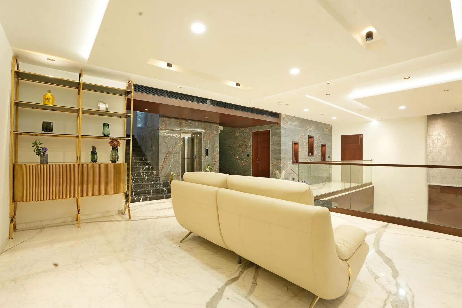 A walls combine light beige paint with grey marble cladding, balancing warmth and coolness. Wooden door frames, dark veneer finishes, and a golden shelving unit add depth, texture, and a touch of elegance.