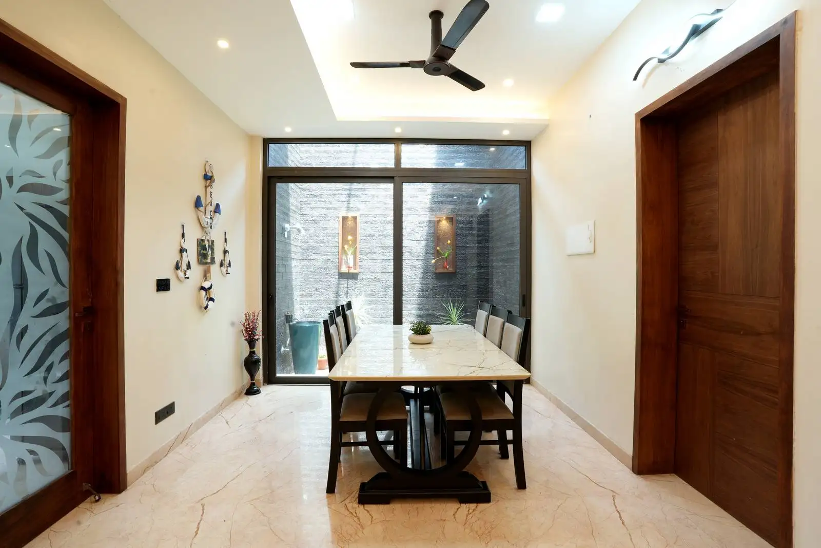 Elegant dining area opens to a serene courtyard through glass panels, blending indoor and outdoor space. Recessed ceiling lighting and clean lines enhance the architectural sophistication.