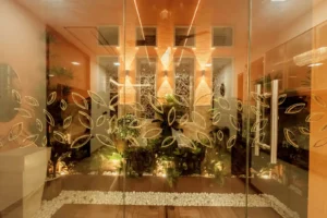 Interior view of a modern room with glass doors decorated with leaf patterns, featuring warm lighting, potted green plants, white pebbles on the floor, and textured walls with small windows.