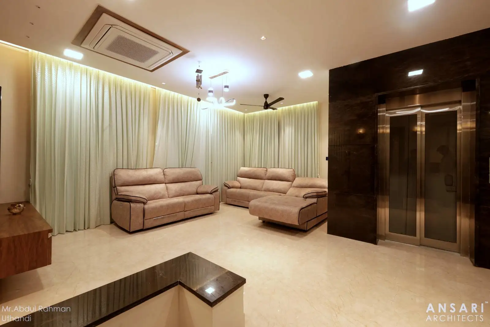 A spacious living room with a minimalist layout, featuring plush sofas for gatherings and a sleek built-in elevator with a glossy black surround that adds both luxury and accessibility.