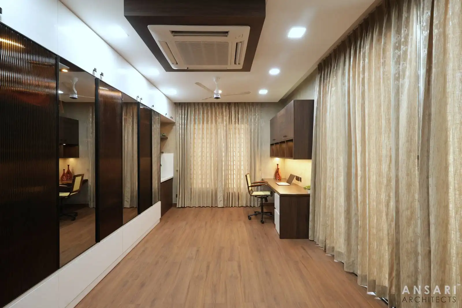 Library room with wooden flooring, glass-fronted cabinets, beige curtains, and a study area with desk and overhead storage.