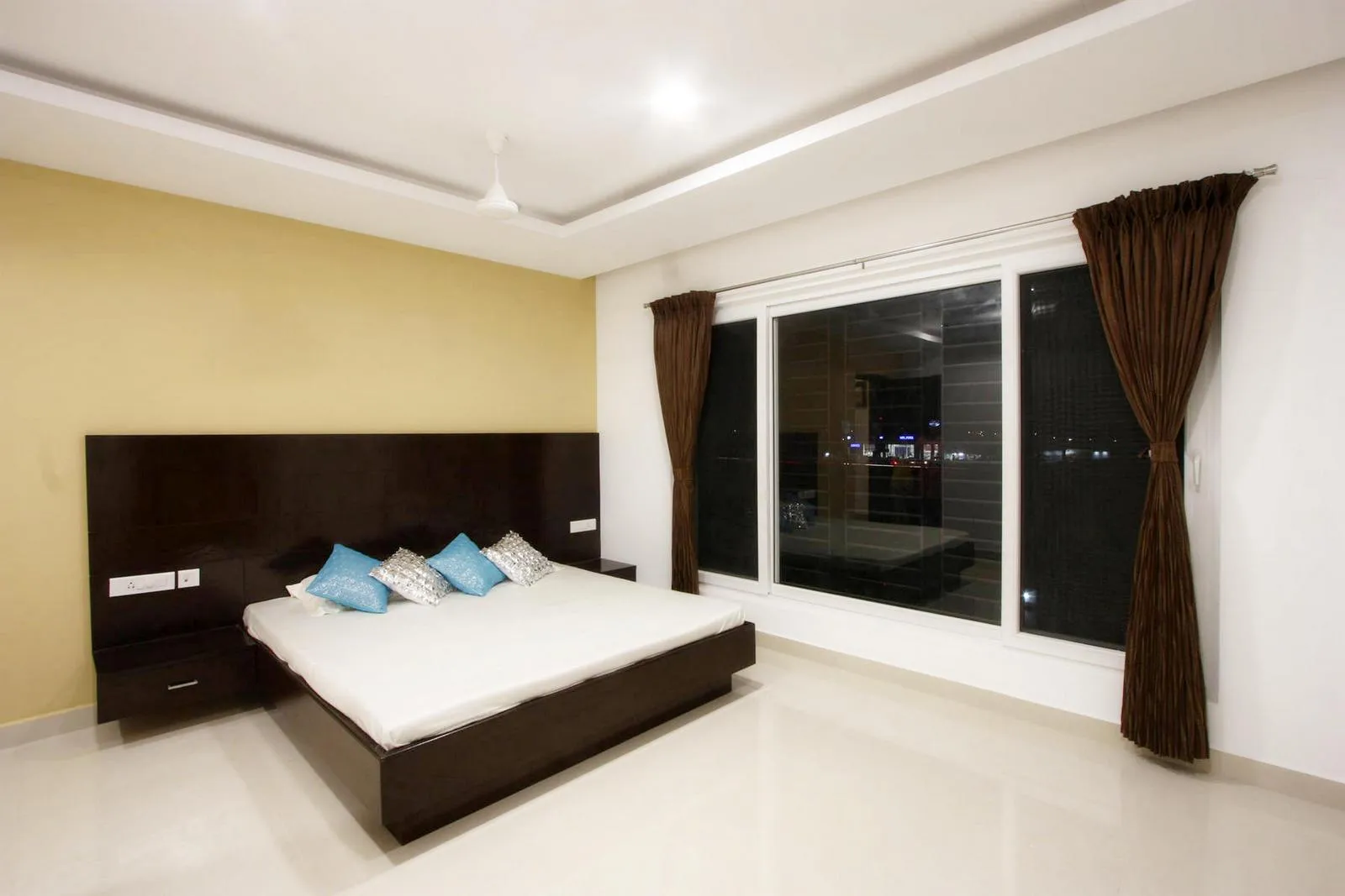 The second-floor bedroom features a rectilinear layout with minimal furnishings, centering the bed against an accent wall. Large sliding glass windows and integrated lighting enhance natural light, views, and a clean, modern aesthetic.