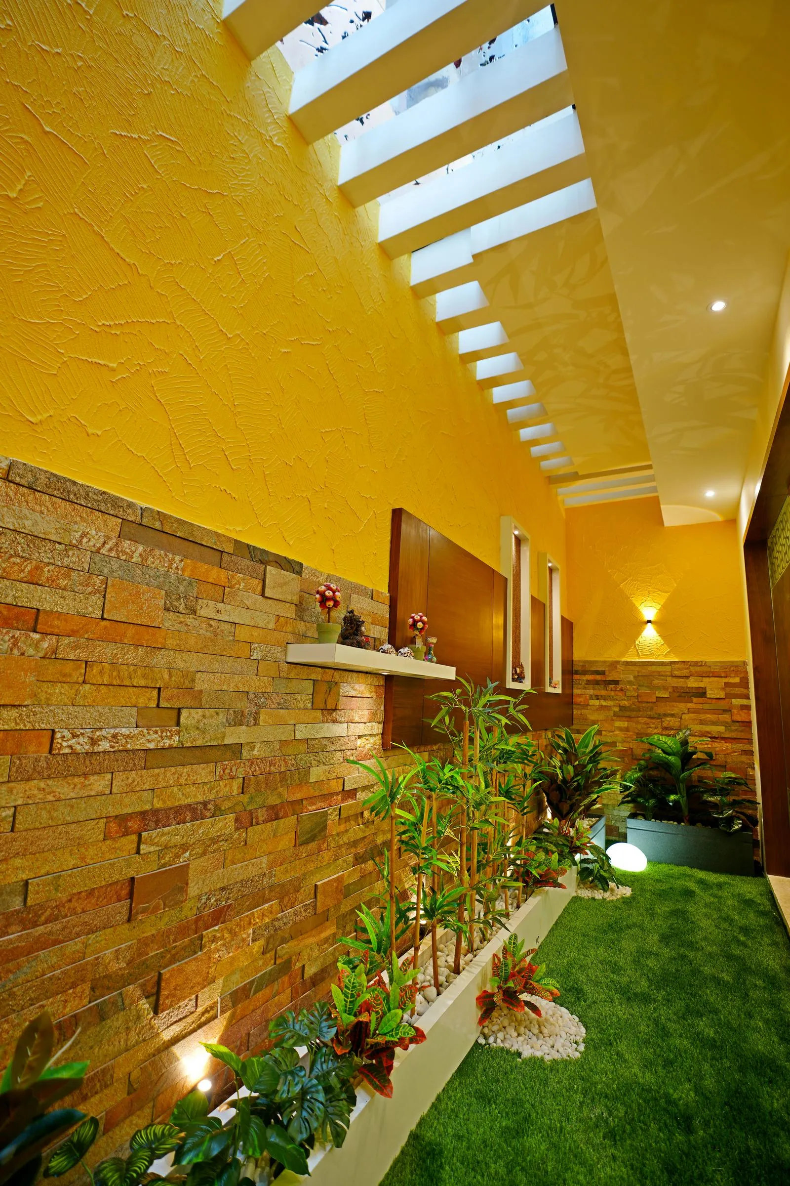 The linear courtyard ( 5'0" x 20'0" sq. ft ) features artificial turf, planter beds, and a skylight with slatted beams, connecting interior spaces with natural light and greenery. Textured yellow walls, stone cladding, and wooden frames create a warm, earthy palette.