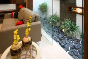 Modern living room with a beige sofa adorned with black and red cushions, facing a glass partition that encloses an indoor garden area featuring smooth black stones, green plants, and a textured wall with a small illuminated shelf containing decorative items, with two brown and beige striped vases holding yellow flowers on a round glass table beside the sofa.