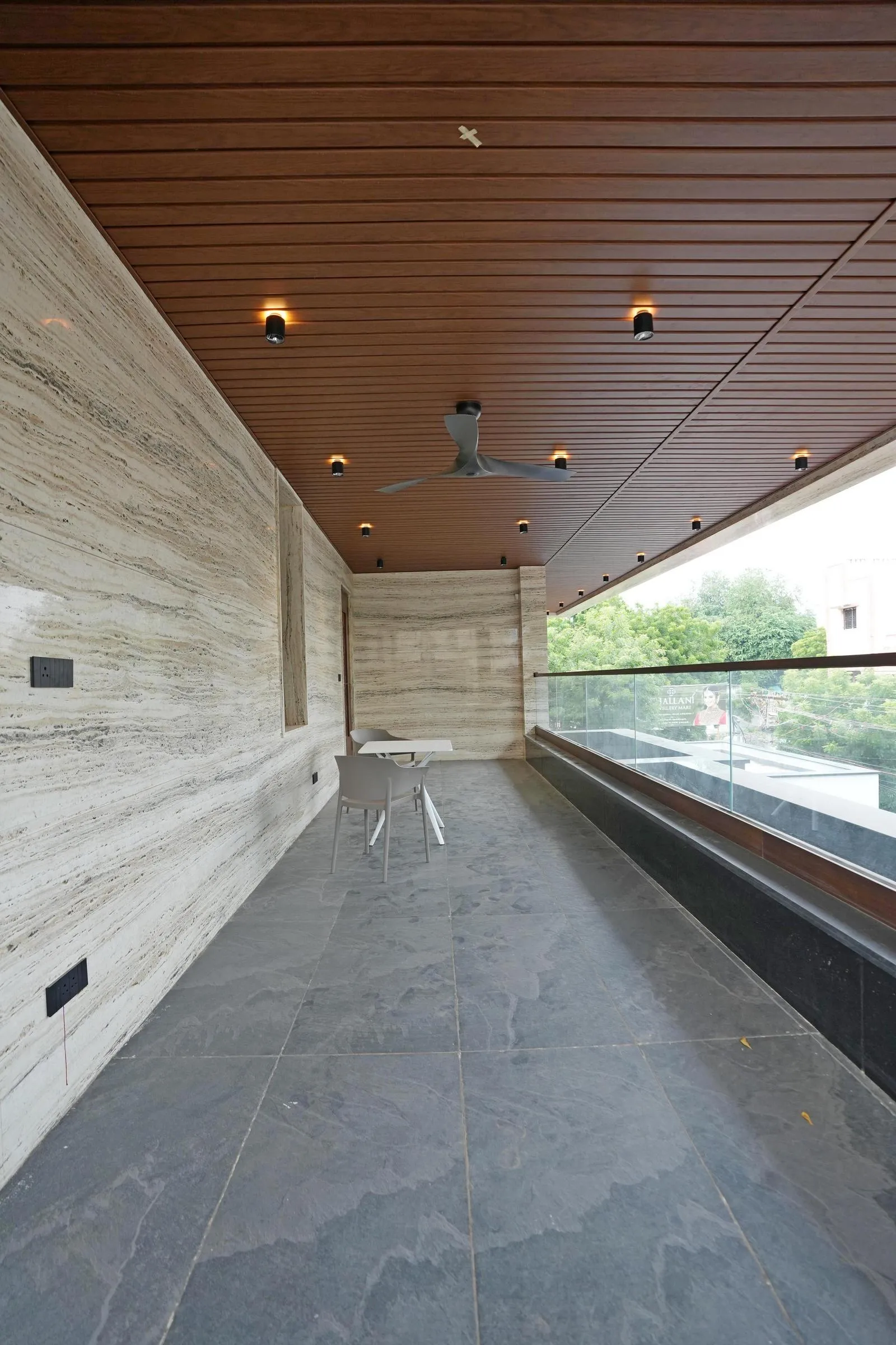 32'0" x 9'0" sit-out balcony extends along the façade, offering a seamless indoor-outdoor connection. Dark grey stone flooring, beige travertine walls, and minimal furniture create a functional, elegant, and open lounging space.