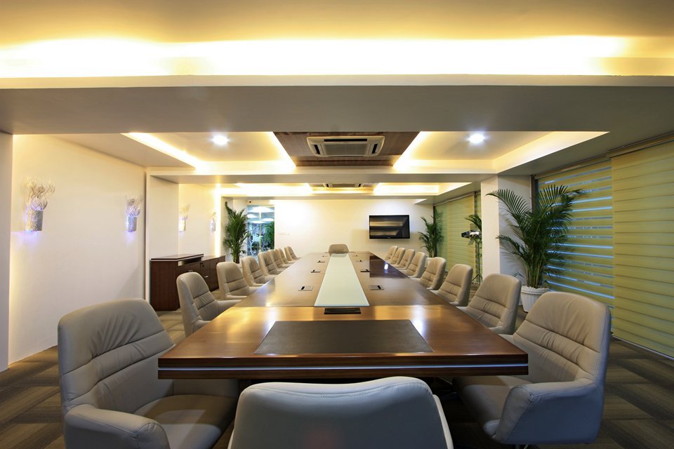Spacious conference room with a long wooden table, soft lighting, green blinds, potted plants, and an integrated screen for professional meetings.