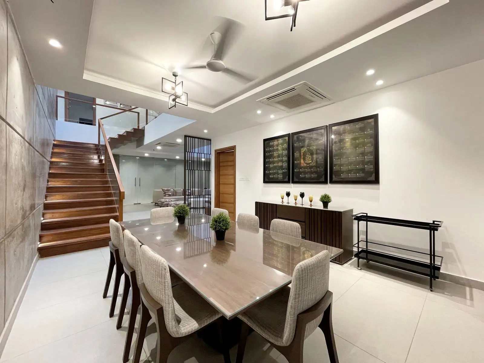 The dining area blends exposed concrete, warm timber, and neutral seating for an open, elegant, and connected space. Pendant lighting and a sculptural staircase enhance flow, light, and architectural interest.