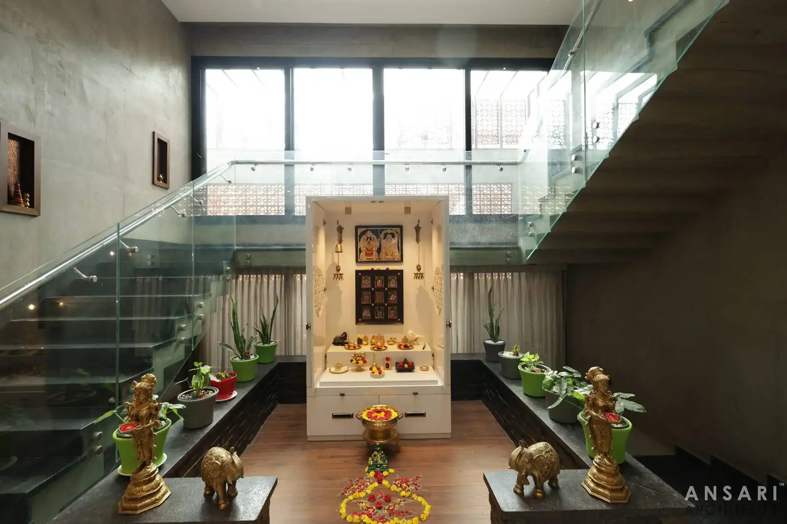 Central puja space with double-height glass, sculptural stairs, and warm textures creates a serene, light-filled courtyard.