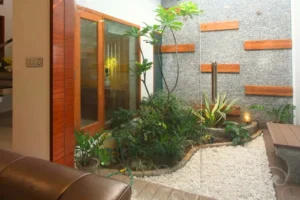 Indoor garden area with green plants, white pebbles, wooden accents on a stone wall, glass door, and a brown leather sofa partially visible.
