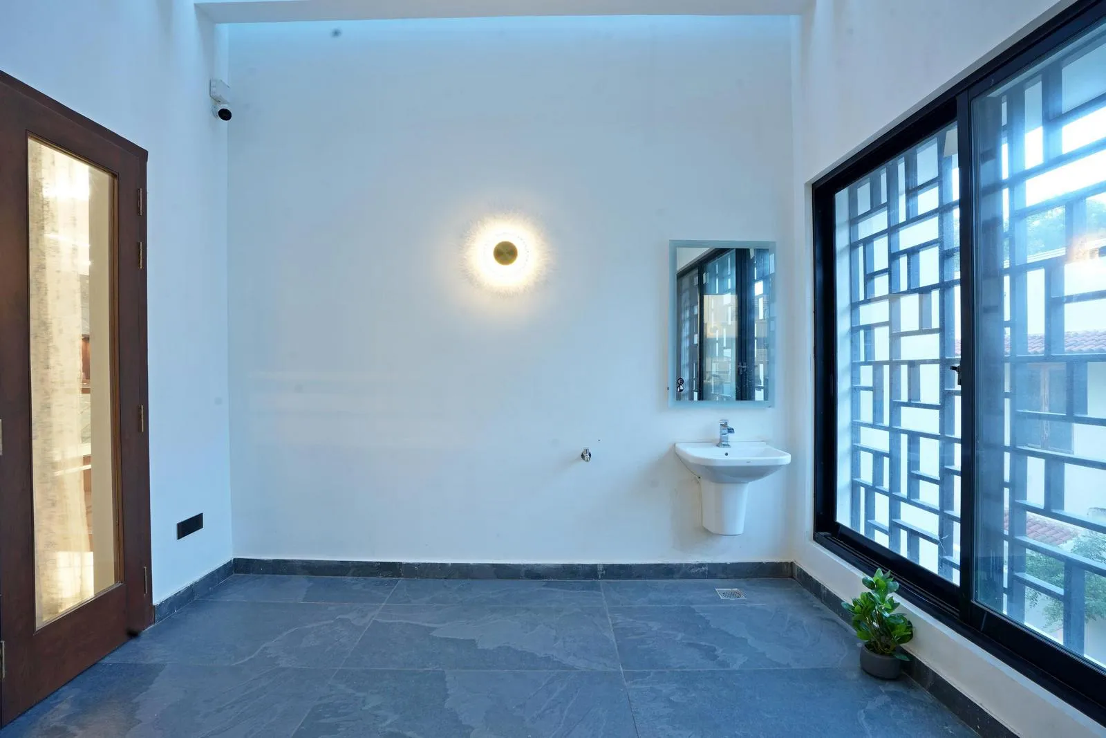 The sit-out area ( 11'6" x 18'3" ) under the pergola features grey tiled flooring, white walls, and a wooden door with a glass panel, creating a clean and minimalist setting. A wall-mounted washbasin, geometric grid window, soft wall lighting, and a potted plant add function, light, and subtle warmth to the space.