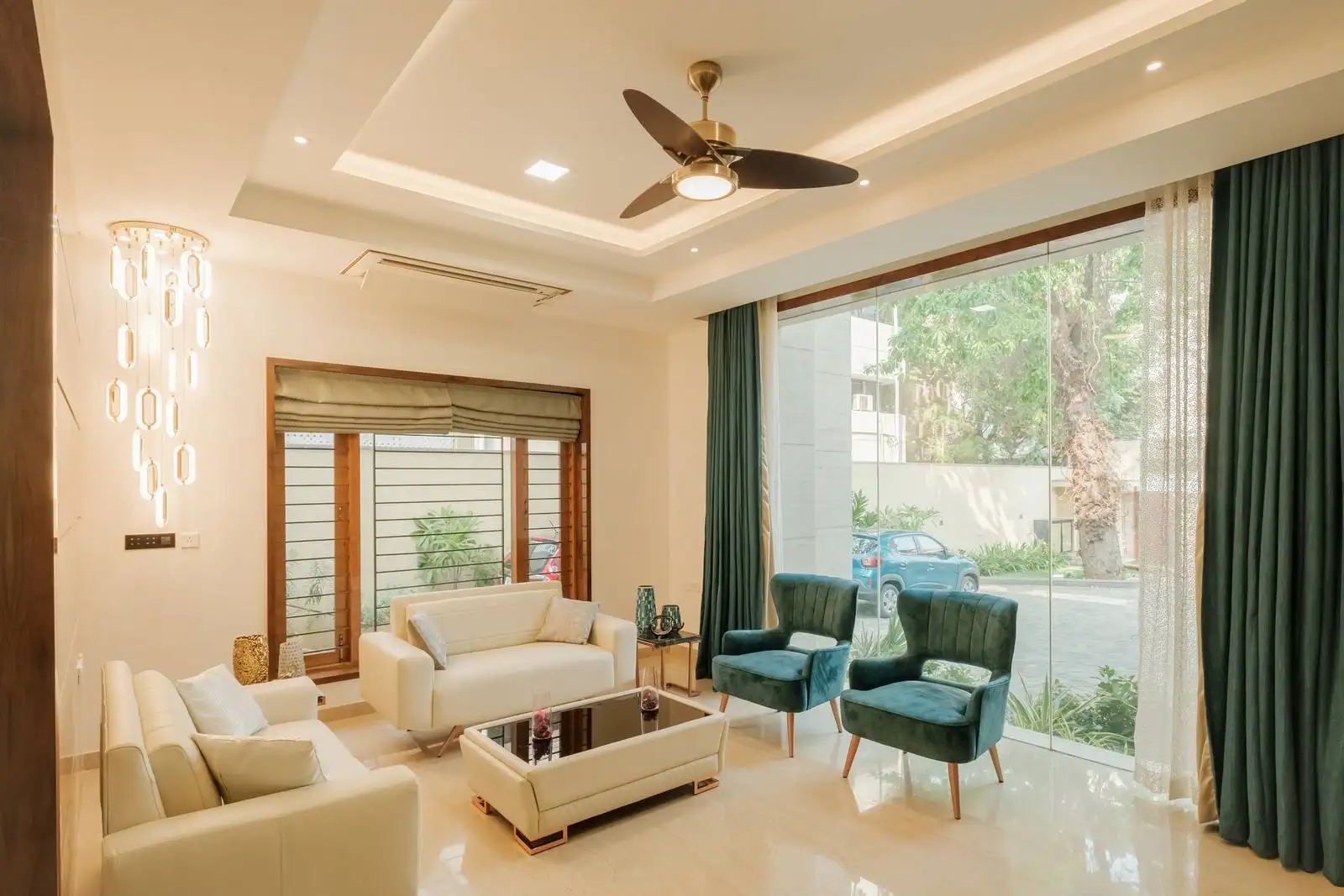 Reception room lighting with chandelier and recessed lights. Comfortable upholstered seating and glass coffee table. Large windows with teal curtains and wooden blinds for light and ventilation control.

