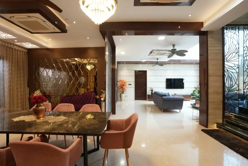 Modern open living and dining area with marble flooring, pink upholstered chairs around a dark marble dining table, a purple tufted sofa near a decorative mirrored wall, a gray sofa facing a wall-mounted TV, ceiling fans, and plants near large windows with artistic grills.