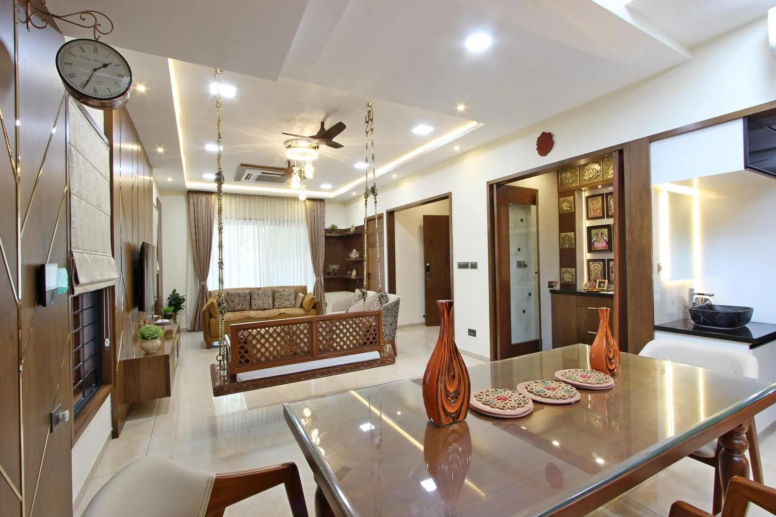 The open-plan design links the living, dining, and puja areas. A wooden swing divides the lounge and dining, while the backlit puja room serves as a spiritual focal point.